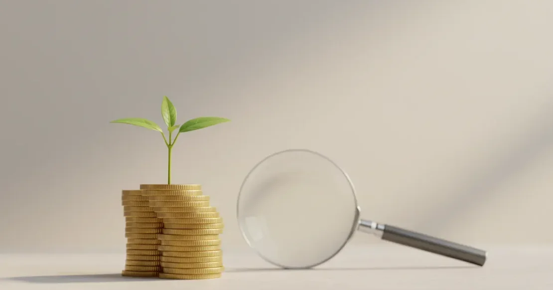 Small green sprout growing from a stack of coins, with a magnifying glass nearby on a soft neutral background.