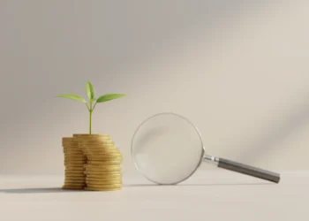 Small green sprout growing from a stack of coins, with a magnifying glass nearby on a soft neutral background.